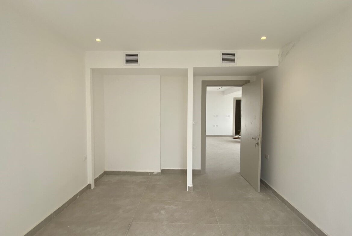 Empty white room with gray tile floor, open doorway to another room and two wall vents above the doorway (unfinished interior).