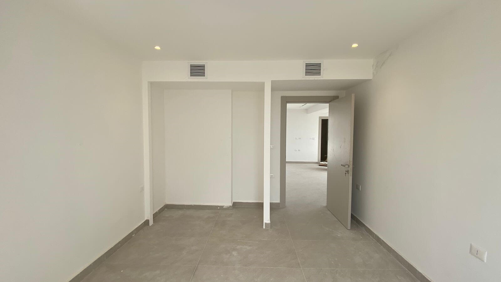 Empty white room with gray tile floor, open doorway to another room and two wall vents above the doorway (unfinished interior).