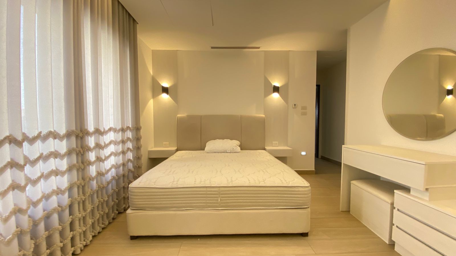 8b80f50c-59d4-4097-9252-1a661077b524 Bright modern bedroom with a padded headboard bed, white nightstands, and sheer vertical curtains on the left wall.