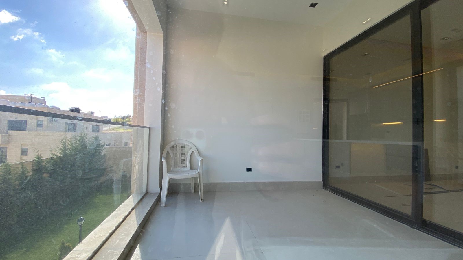 8ce68ee0-18d1-4217-8a0e-1bb2e7e788b6 Enclosed balcony with glass walls and railing, a single white plastic chair, and a view of a sunny apartment complex outside.