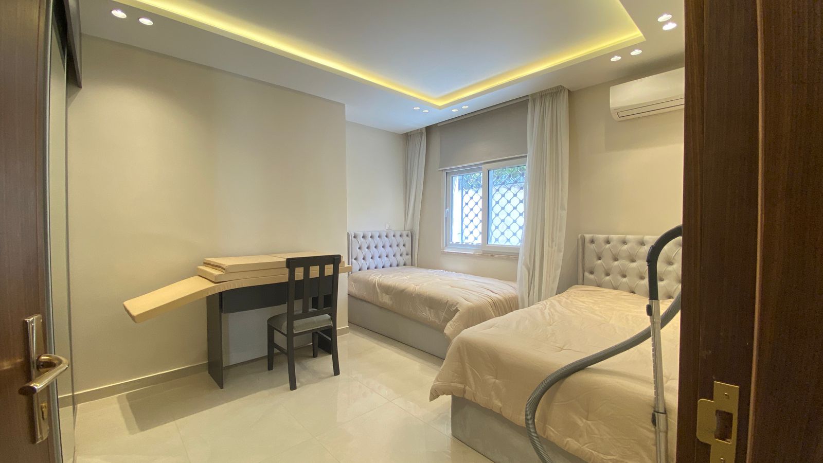 8d5f0bb3-aee0-4b39-8e42-2b12d32743ed Bright contemporary bedroom with two twin beds and tufted headboards, a small desk and chair, and a large window with light curtains.