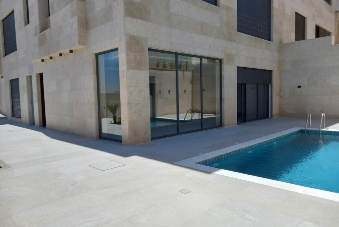 Modern beige stone building with large glass doors overlooking a blue rectangular pool and metal ladder, outdoor patio in bright sunlight