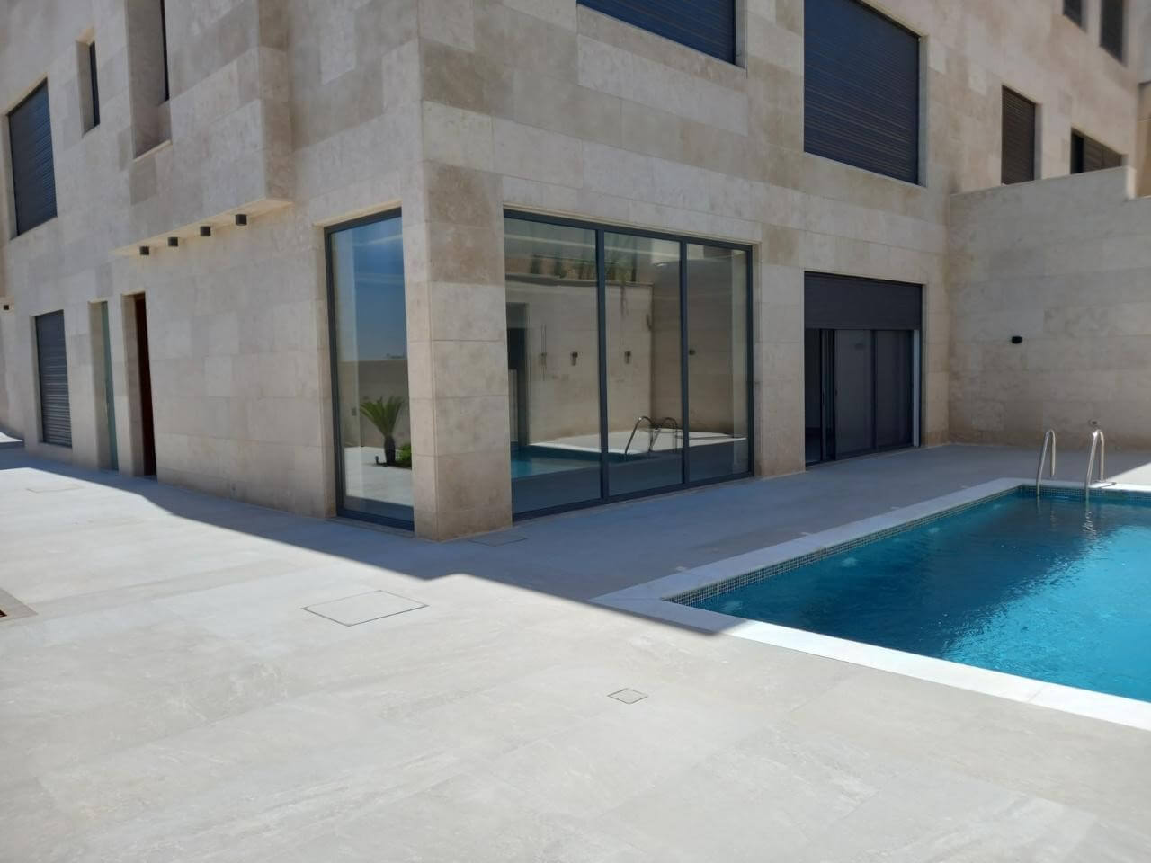 8dc47433-4f43-4f91-b2e0-ca8ccb326e32 Modern beige stone building with large glass doors overlooking a blue rectangular pool and metal ladder, outdoor patio in bright sunlight