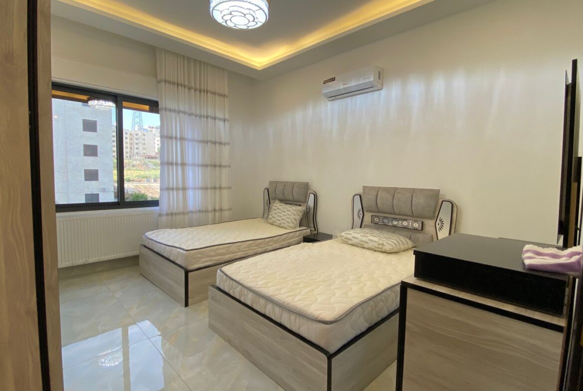 Twin bedroom with two single beds, grey headboards, and a large window overlooking buildings outside.