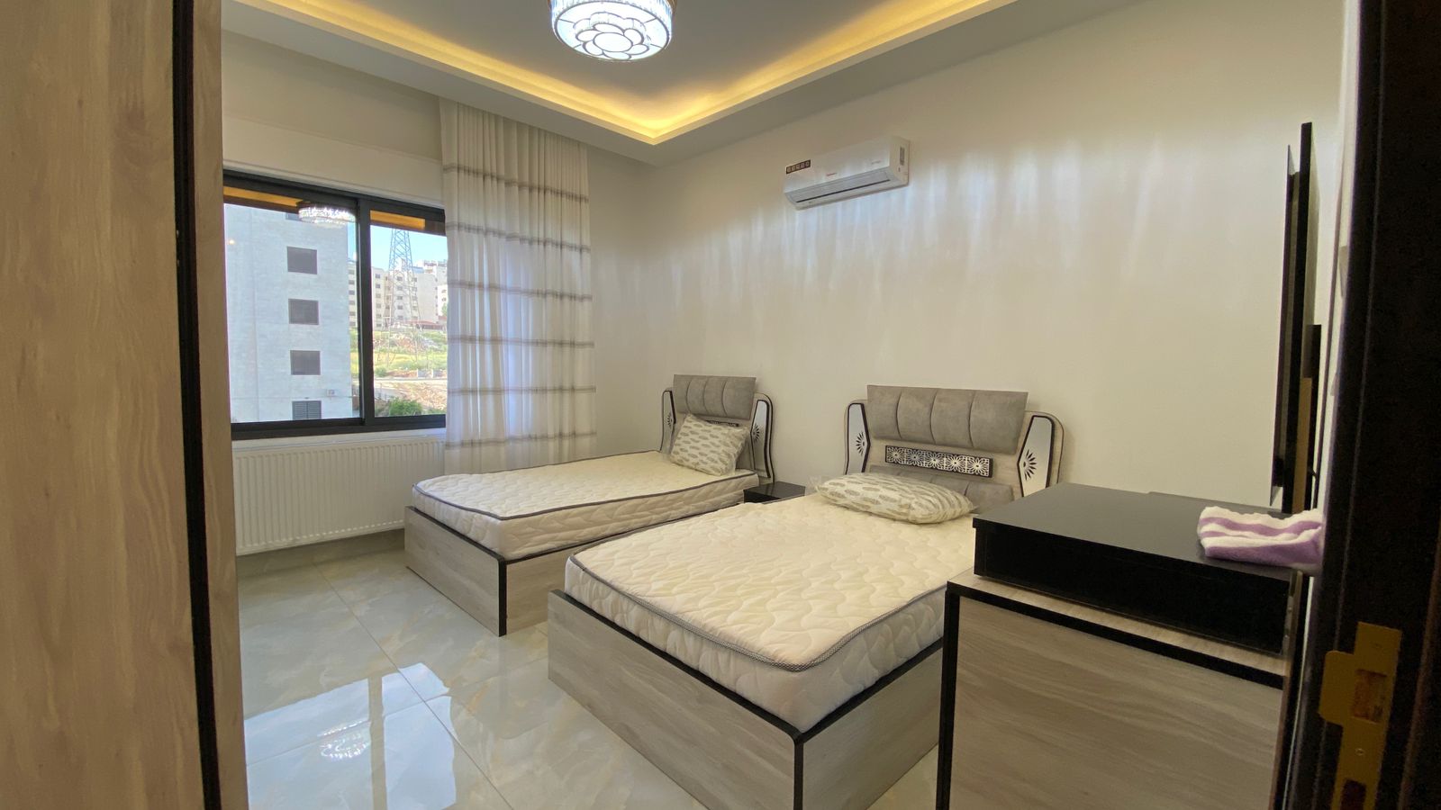 Twin bedroom with two single beds, grey headboards, and a large window overlooking buildings outside.
