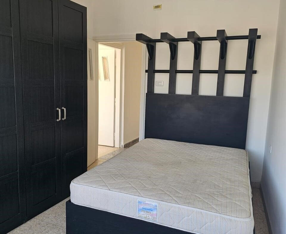 8e23c879-989e-4d19-bf47-a59bac489030 Bedroom with a black wooden bed frame and a high headboard featuring vertical slats and hooks; mattress on the bed, and a tall black wardrobe along the left wall.