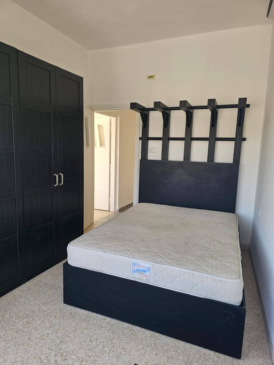 8e23c879-989e-4d19-bf47-a59bac489030 Bedroom with a black wooden bed frame and a high headboard featuring vertical slats and hooks; mattress on the bed, and a tall black wardrobe along the left wall.