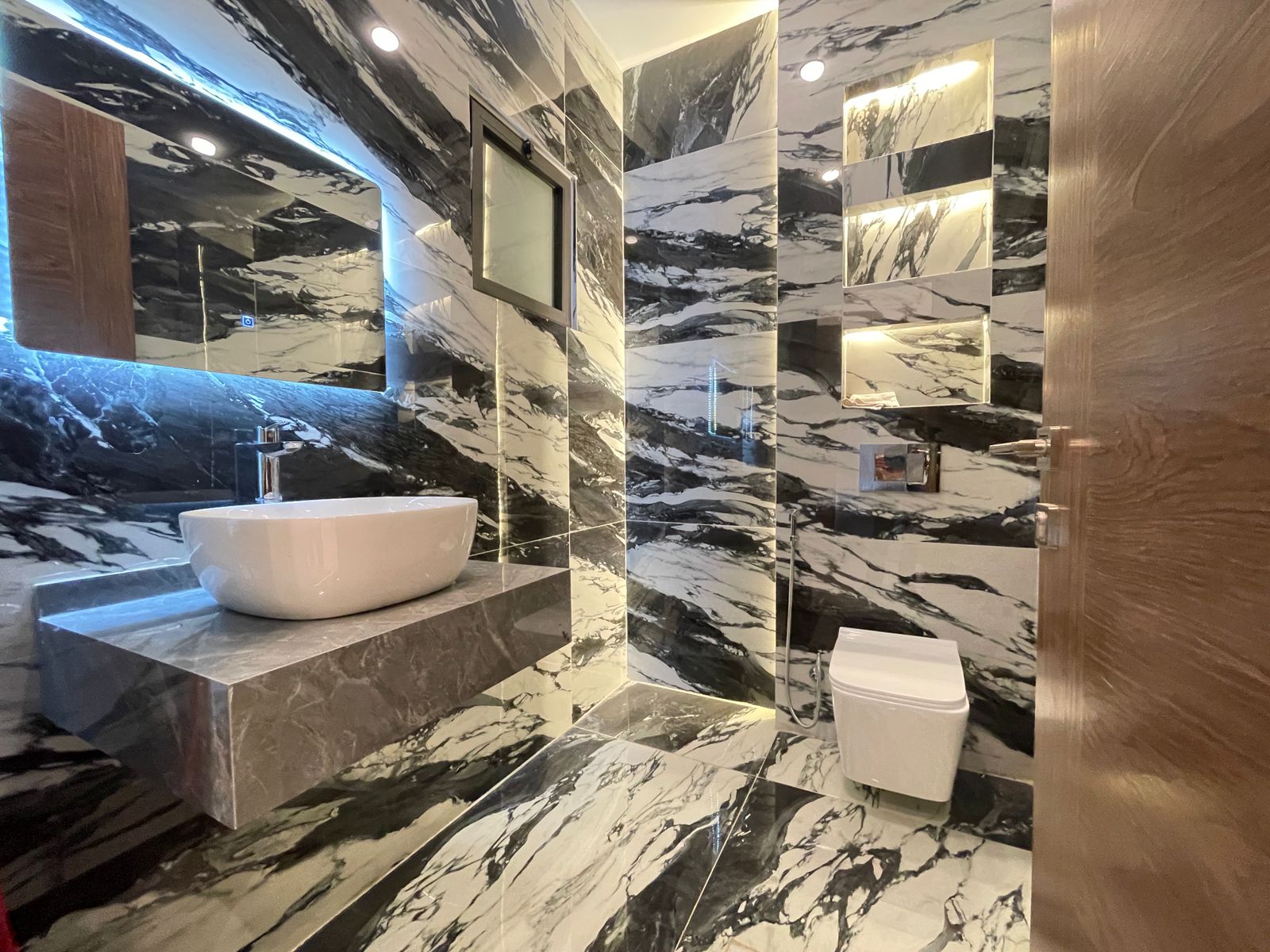 8e417a86-04a4-4456-aa3c-04438cc4f2b7 Luxurious marble bathroom with black and white veined walls, a white vessel sink on a marble counter, and a large mirrored wall reflecting blue LED lighting.