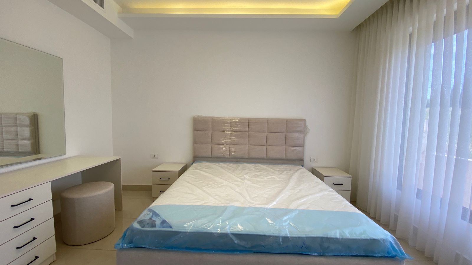 8ebec9c1-4e9d-493b-9de3-611803d6e584 Bright, minimalist bedroom with a padded headboard bed, white nightstands, and a large mirror near a white dresser and stool.