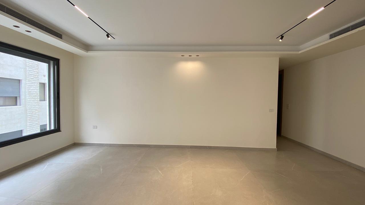 8ee97fb7-4438-493c-866a-b7ae658ffbcd Empty modern living room with a large left window, beige walls, and track lighting across the ceiling.