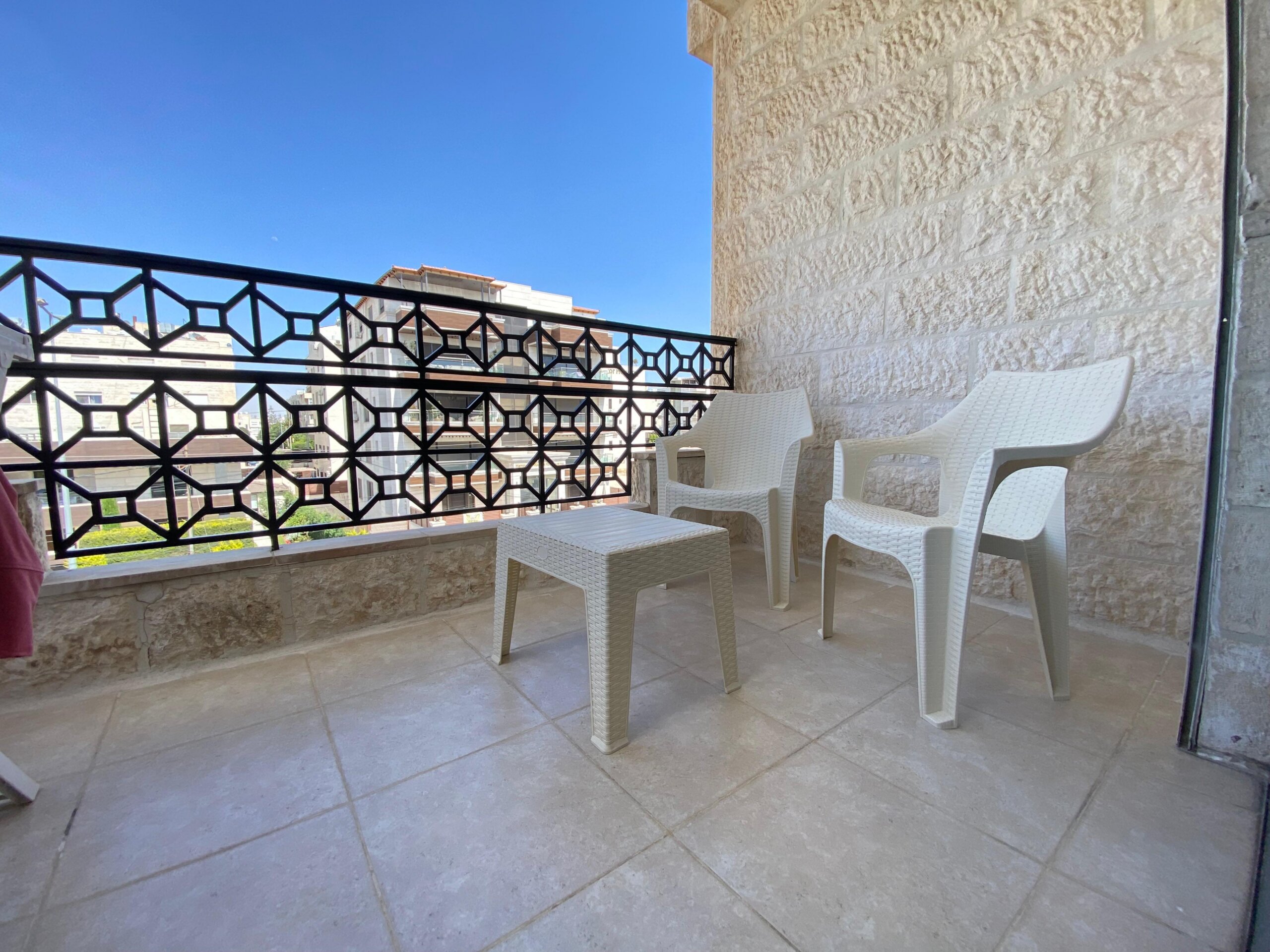 8ef04e4b-5683-4297-a7ea-6e36e942fd1c Balcony with white plastic chairs along a beige stone wall and a black geometric railing against a clear blue sky.