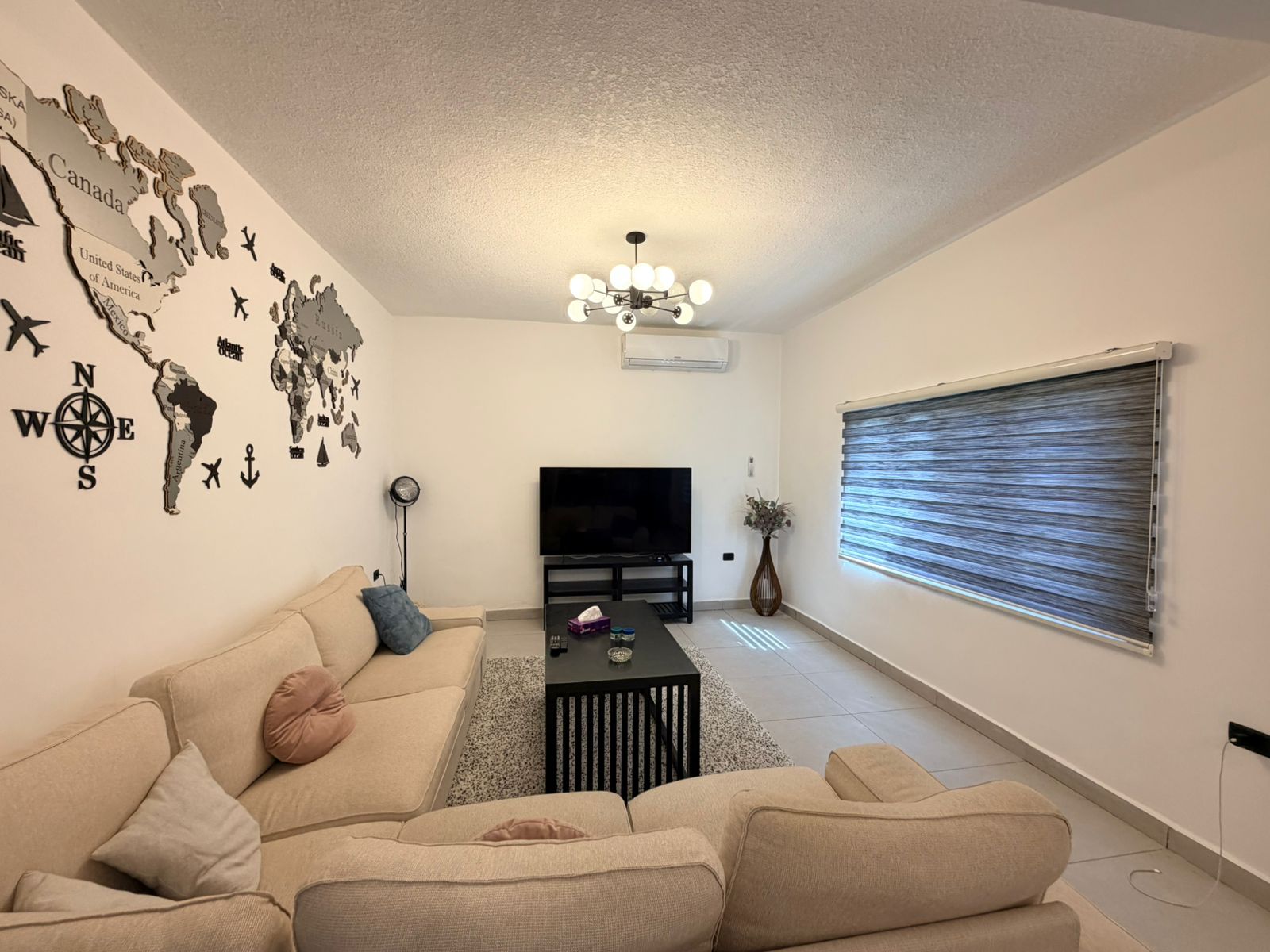8f222072-b562-4466-8257-3ab1f1a42900 Cozy living room with a beige sectional, black coffee table, travel/world map wall art, and a TV on a stand under a modern chandelier.
