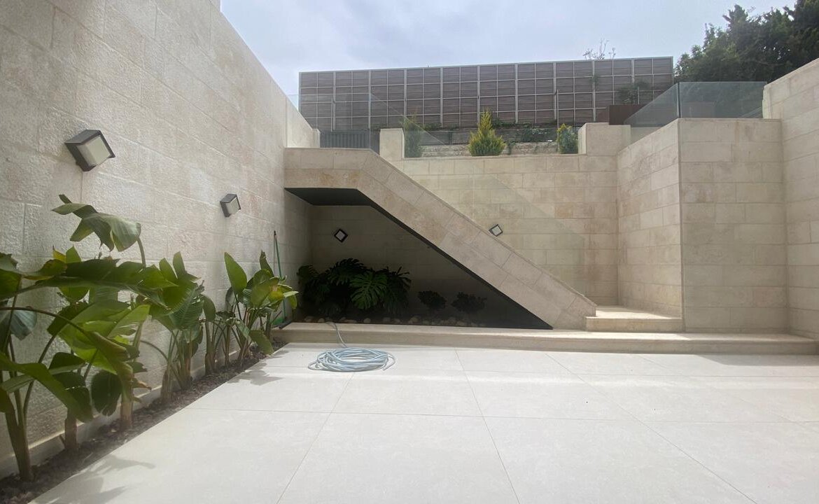 A sunlit beige stone courtyard with a wide staircase under an angled wall, plants along the left wall, and a coiled hose on the tiled ground.