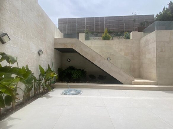 A sunlit beige stone courtyard with a wide staircase under an angled wall, plants along the left wall, and a coiled hose on the tiled ground.