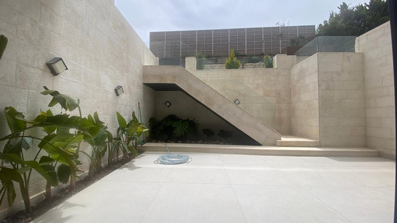 8f45a6d6-653c-49c1-8fc5-4520170d9a0e A sunlit beige stone courtyard with a wide staircase under an angled wall, plants along the left wall, and a coiled hose on the tiled ground.