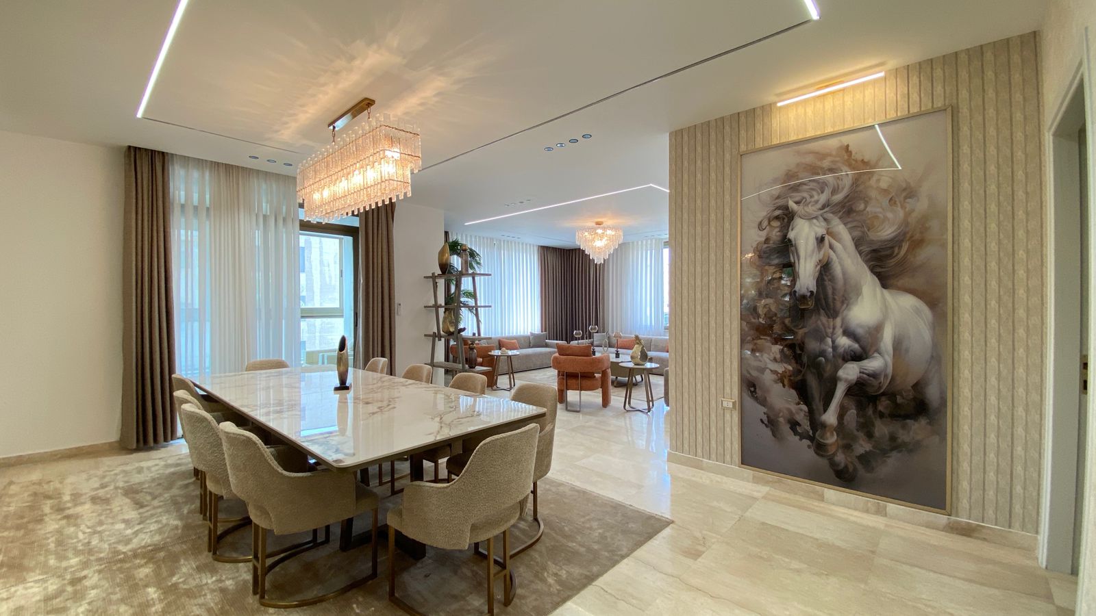 8fb45fda-c0e9-4d02-a678-a5e1cf9cb8d1 Open-plan dining and living area with a marble dining table, beige upholstered chairs, and crystal chandeliers; a large horse artwork sits on a textured wall to the right.