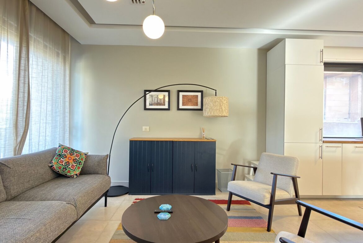Living room with a gray sofa, colorful patterned cushion, round coffee table, and a large arc floor lamp beside a navy cabinet against a pale wall.