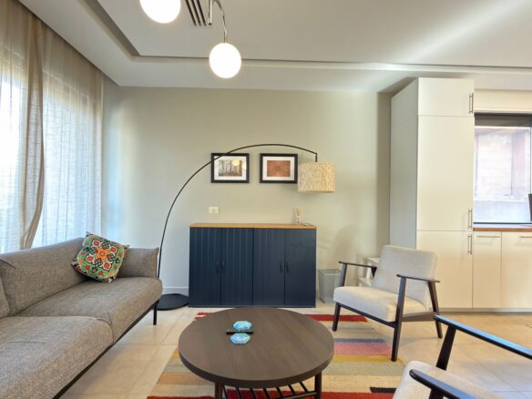 Living room with a gray sofa, colorful patterned cushion, round coffee table, and a large arc floor lamp beside a navy cabinet against a pale wall.