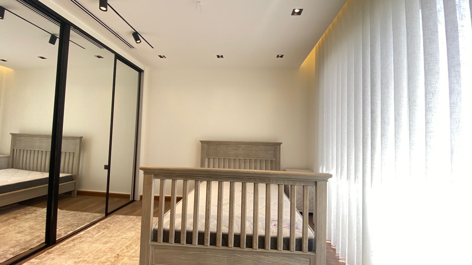 9154571a-3024-41bf-aec4-3f505c8fa583 Light-filled bedroom with light wood furniture, a wooden crib in the foreground, and a large mirrored wardrobe on the left. Tall vertical blinds cover the right wall.