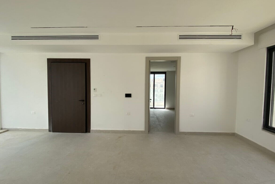 Empty bright room under construction with large windows on both sides, dark wooden door to the left, and a doorway leading to another sunlit space.
