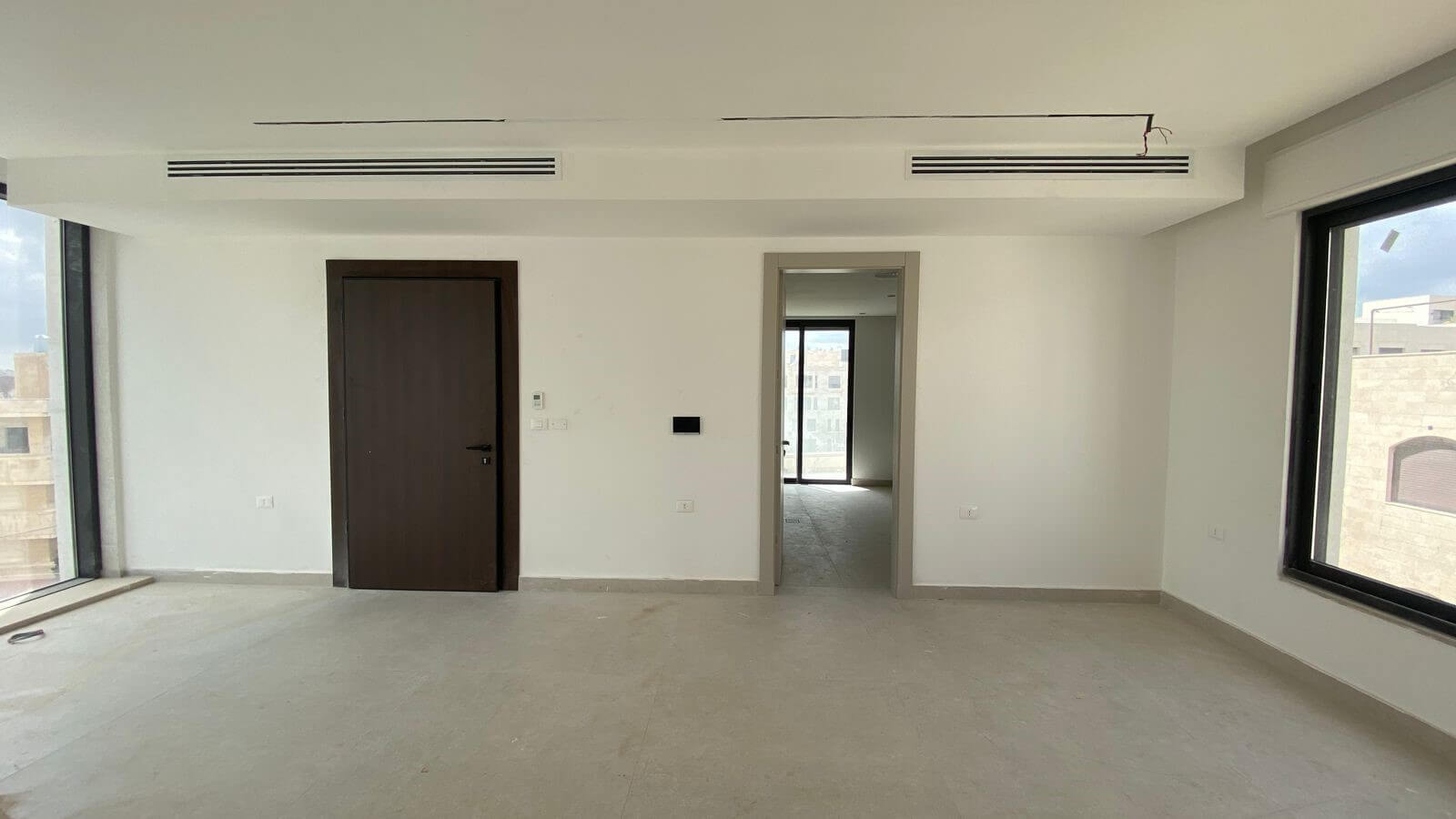 917c5b34-8798-4253-83b8-20e80cb93ae8 Empty bright room under construction with large windows on both sides, dark wooden door to the left, and a doorway leading to another sunlit space.
