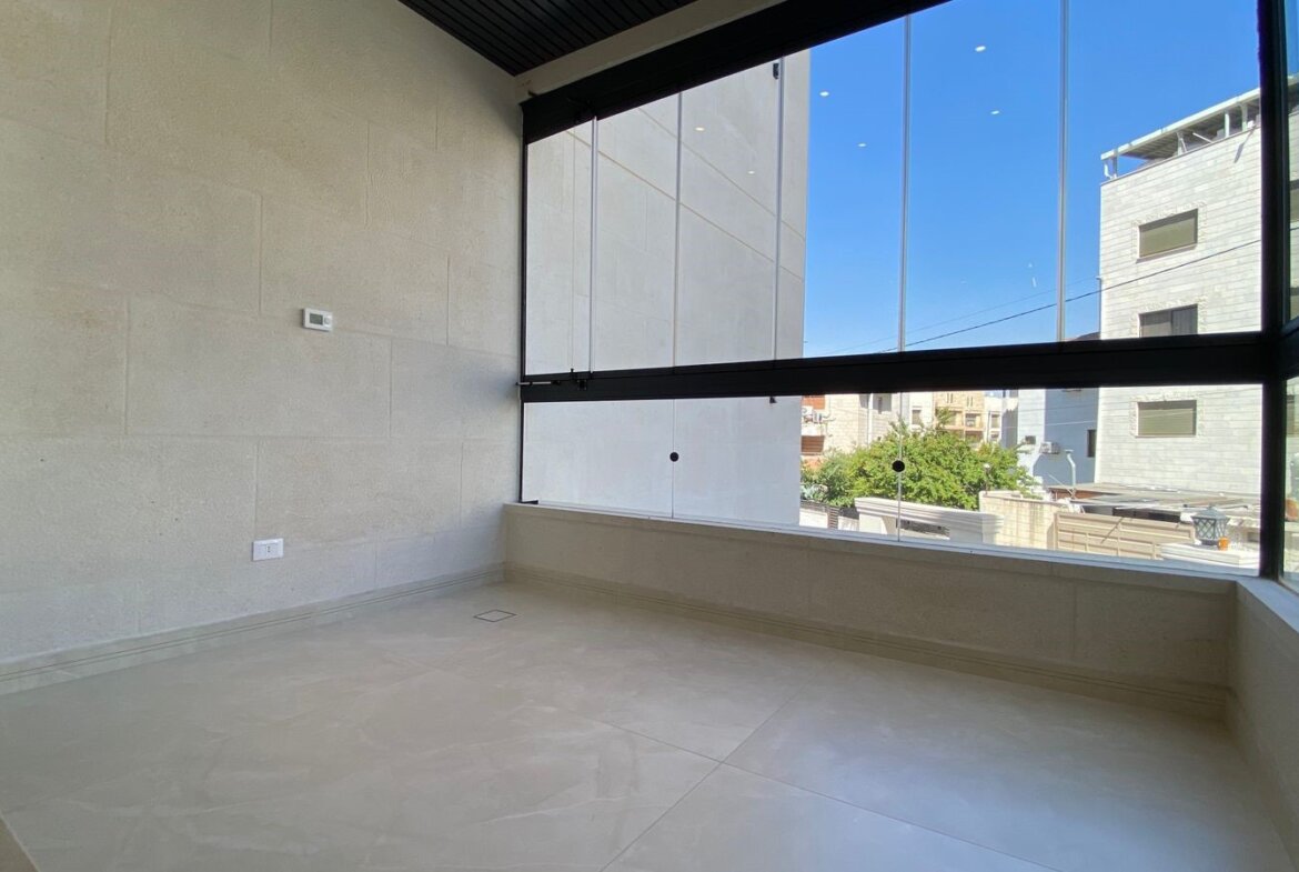 Empty glass-walled balcony with beige stone walls, white tiled floor, and a view of urban buildings on a sunny day