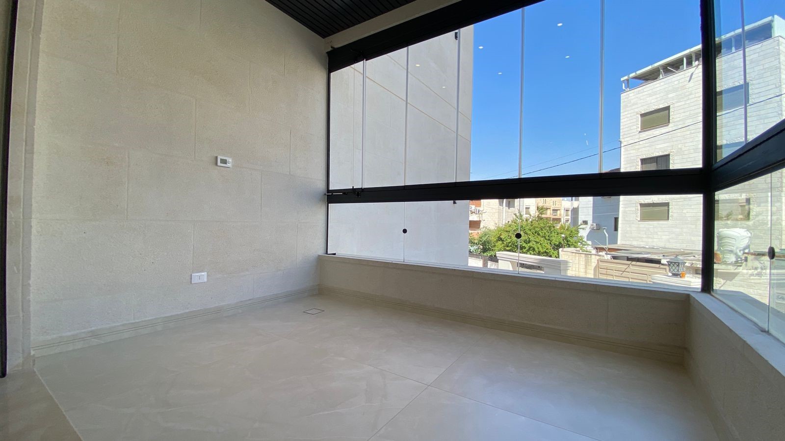 92031dc6-1f41-42ef-82f7-b910a8bdc43e Empty glass-walled balcony with beige stone walls, white tiled floor, and a view of urban buildings on a sunny day