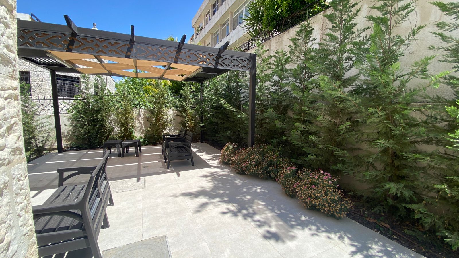925cdddf-7684-4f4a-8b9a-6a7b95760a9c Patio area with black seating under a decorative pergola, surrounded by tall green hedges and a stone wall on a sunny day