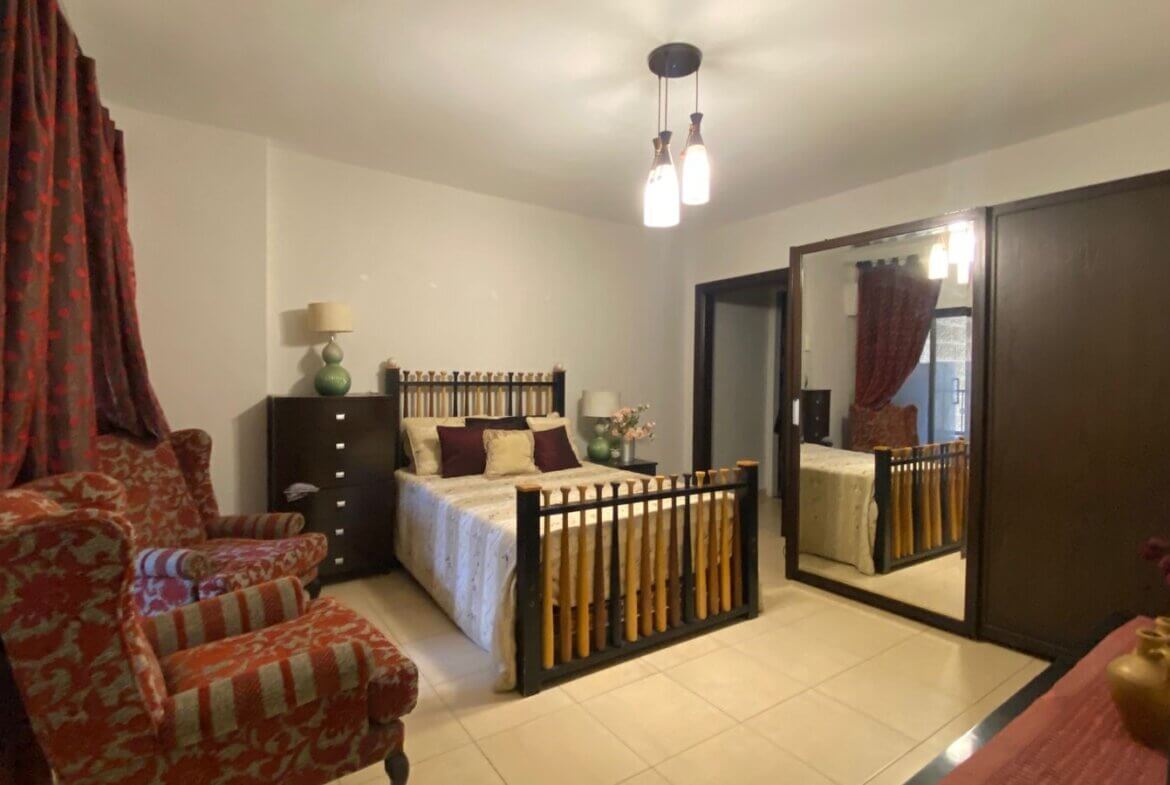 93b0837c-db36-436a-b1da-1229f24e8953 Cozy bedroom with a wooden double bed, matching dressers, green lamps, and red patterned curtains with two matching armchairs nearby.
