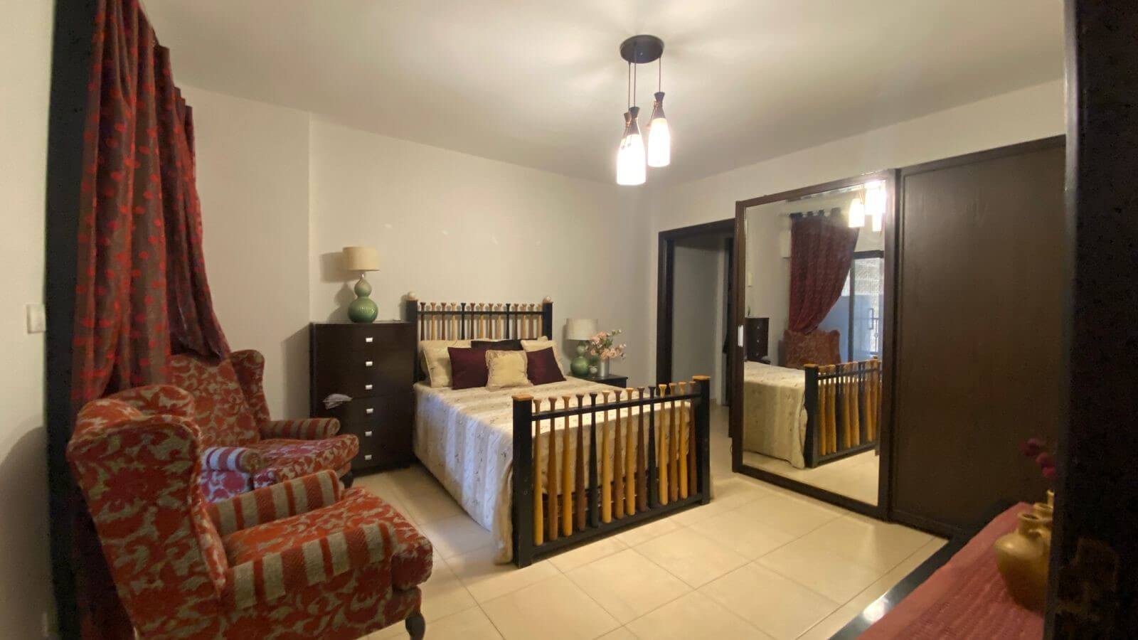 93b0837c-db36-436a-b1da-1229f24e8953 Cozy bedroom with a wooden double bed, matching dressers, green lamps, and red patterned curtains with two matching armchairs nearby.