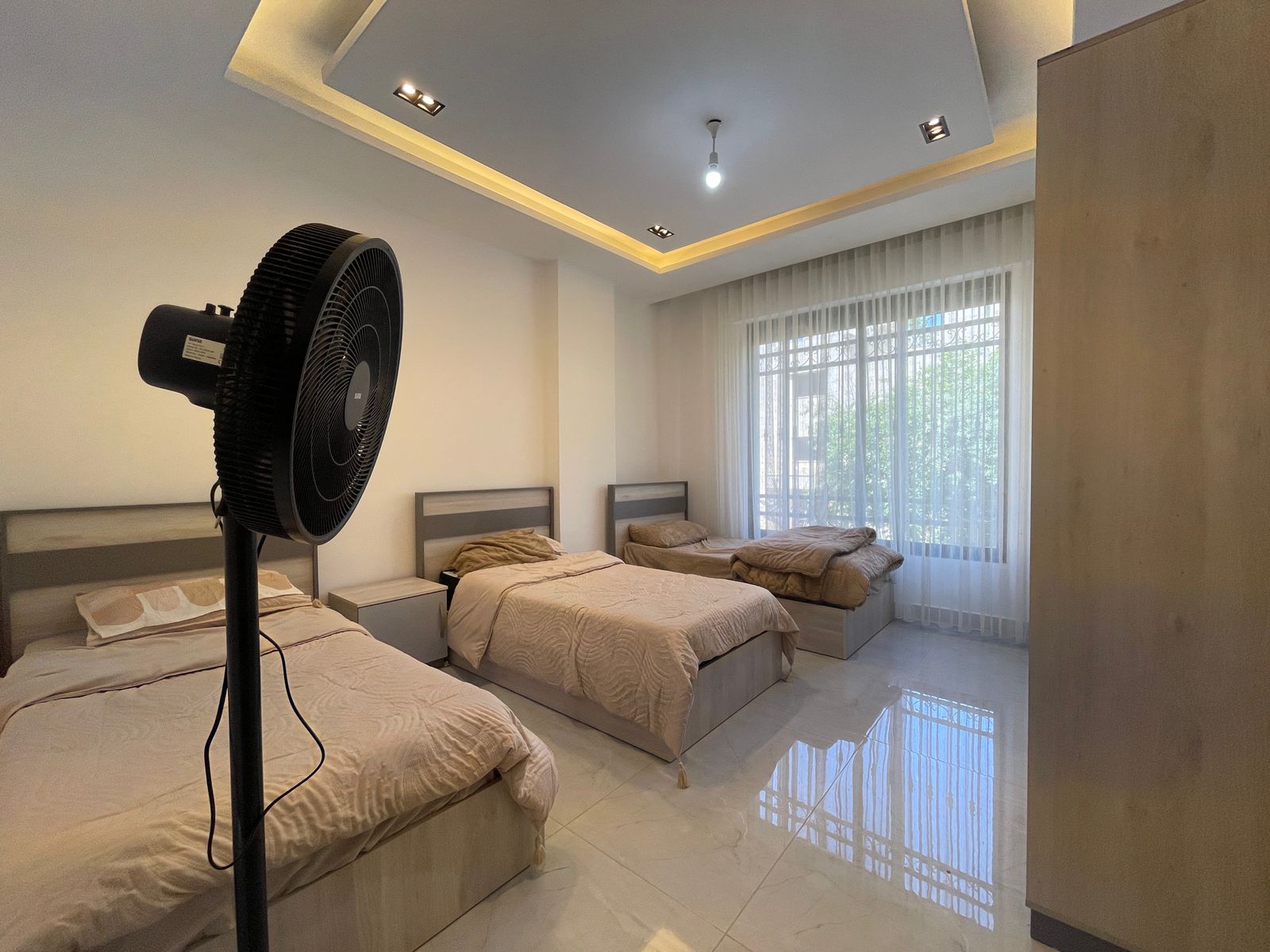 952c586a-b159-4fbf-bf5a-d8a601fd51e3 1 Bedroom with three single beds and a large pedestal fan in the foreground, neutral beige bedding and modern bedside tables nearby.