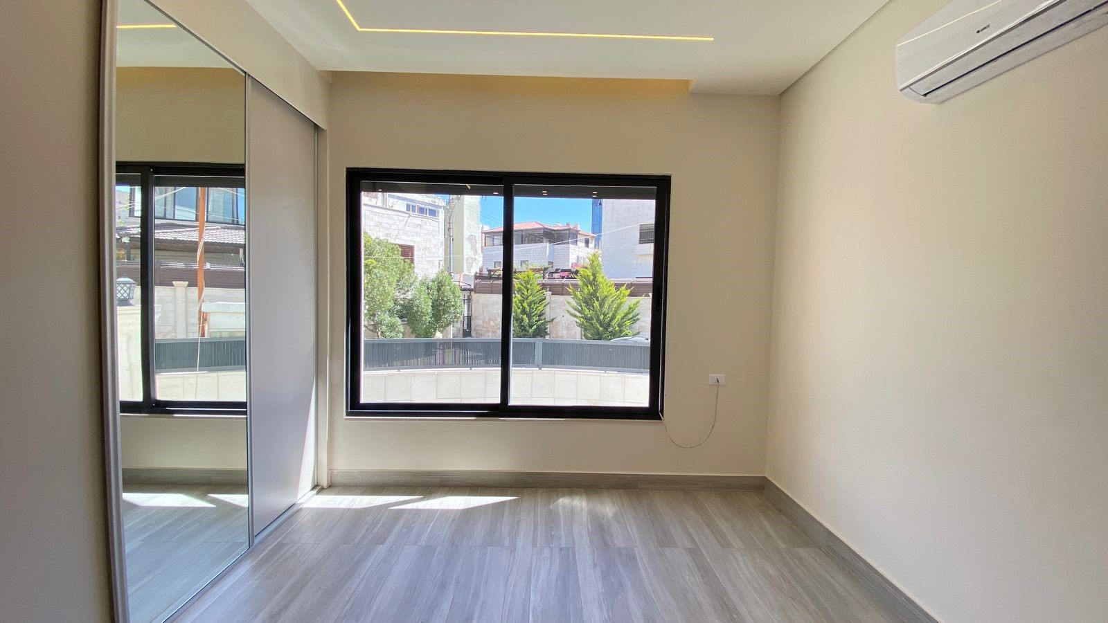 95a7cb31-c6fb-4369-be6f-0cfa2ce7f9e3 Empty modern bedroom with a large window, light wood flooring, and a mirrored sliding-door wardrobe.