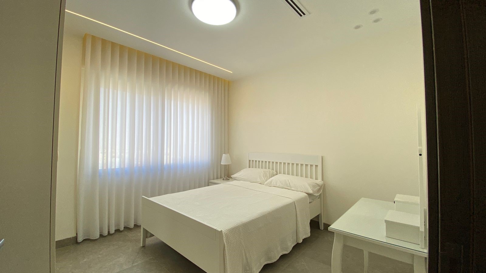 95f679e9-af7b-4290-8b33-412fd94a90f1 Neutral white bedroom with a double bed, white bedding and headboard, nightstand with lamp, and sheer curtains along a large window.