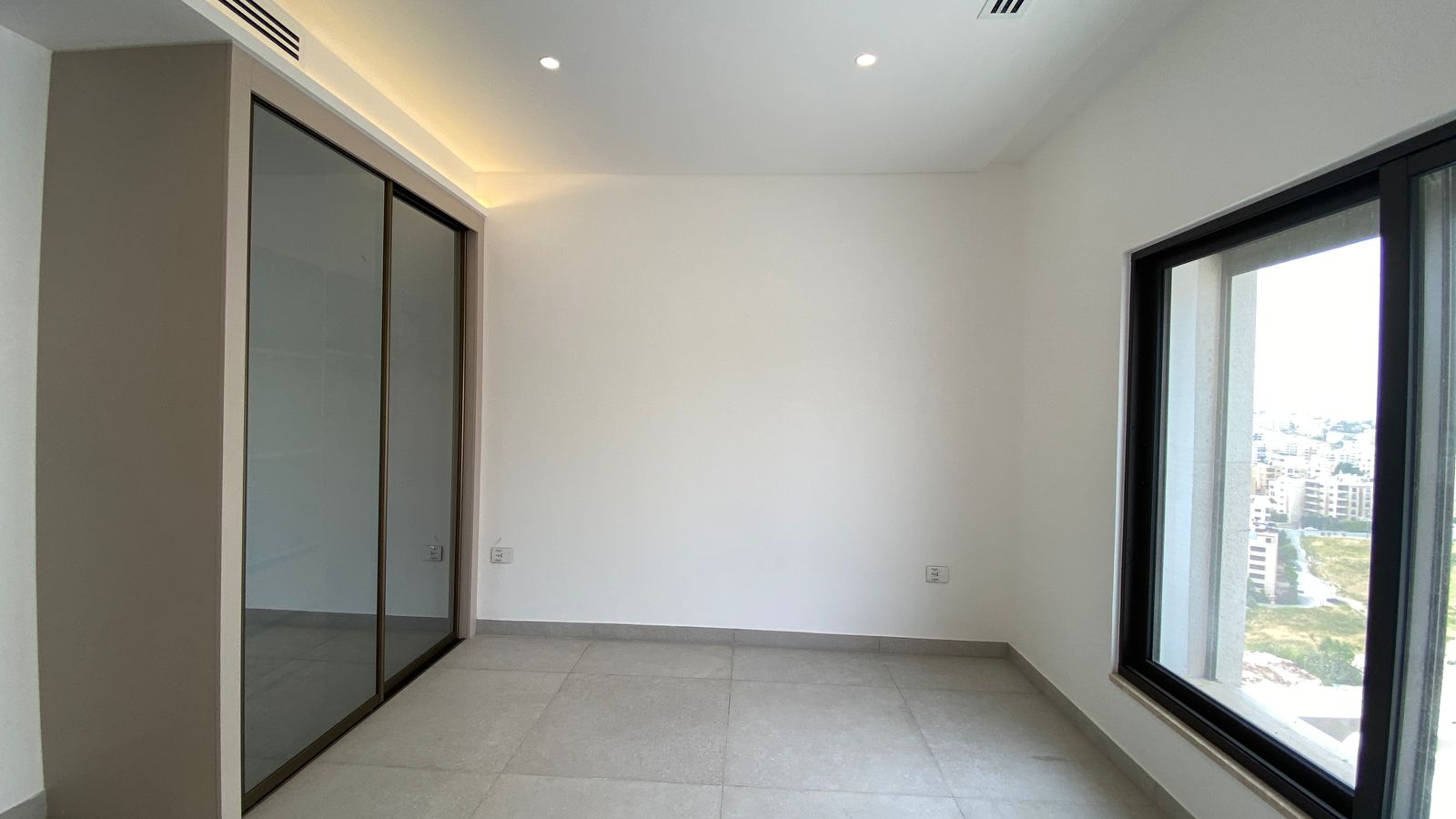 96406915-afb7-4df5-b2f0-1a64a462236c Empty modern room with a large window to the city, light gray tiled floor, and a sliding glass wardrobe on the left; recessed ceiling lights above.