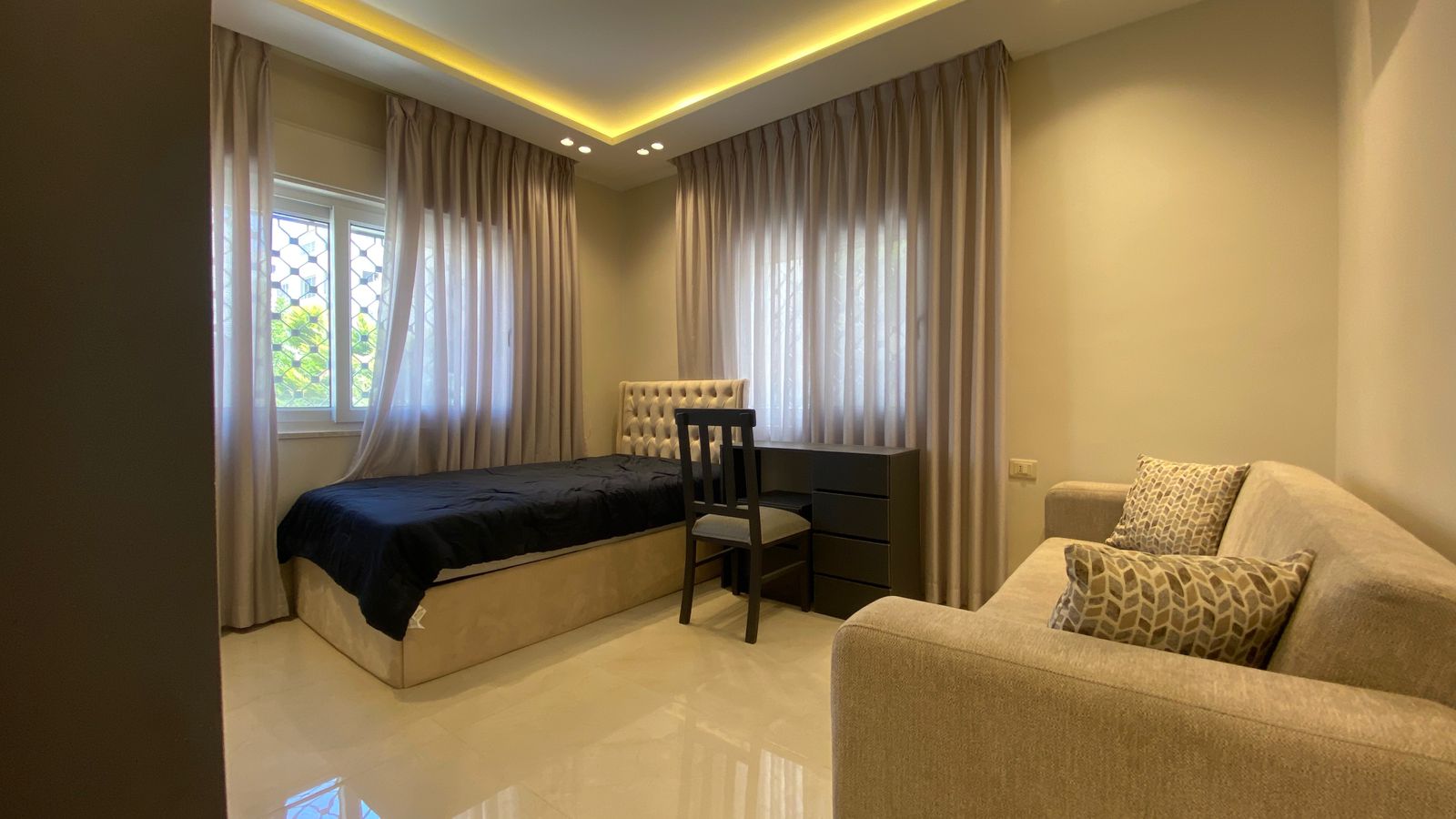 96d1bc92-396b-4dd0-9251-f347e92a4b8c Modern bedroom with a low bed and tufted headboard, a dark desk and chair, and a beige sofa under warm ceiling lighting.
