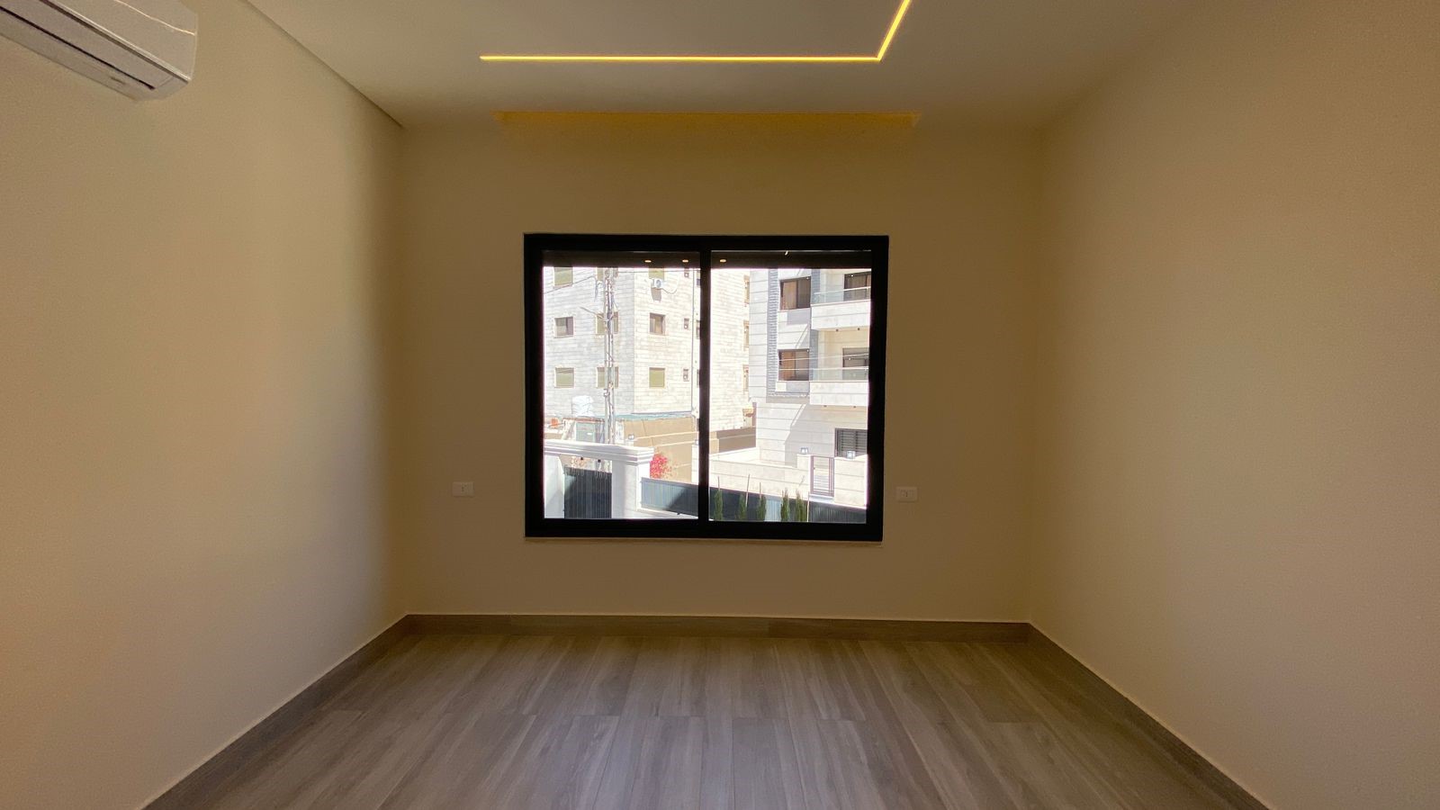 96e3a972-c24f-4205-a52c-cf1c8476503d Empty rectangular room with beige walls, light wood floor, and a large window facing an urban exterior; wall AC unit on left and a yellow ceiling light feature