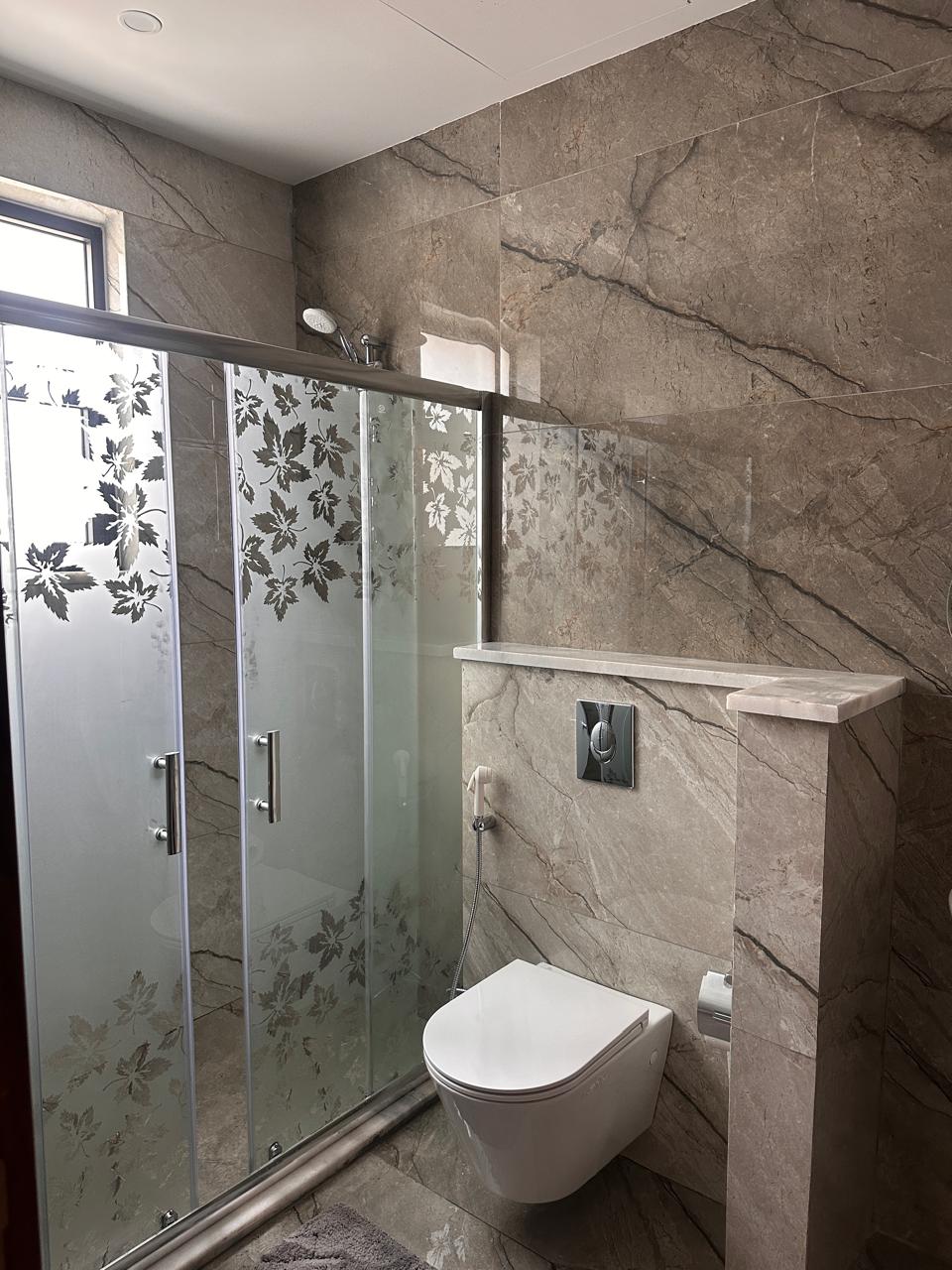970163bb-2942-409d-962f-2724ba7ccd72 Modern bathroom featuring a glass shower enclosure with frosted leaf pattern, a wall-mounted toilet with a chrome flush plate, and marble-tiled walls.