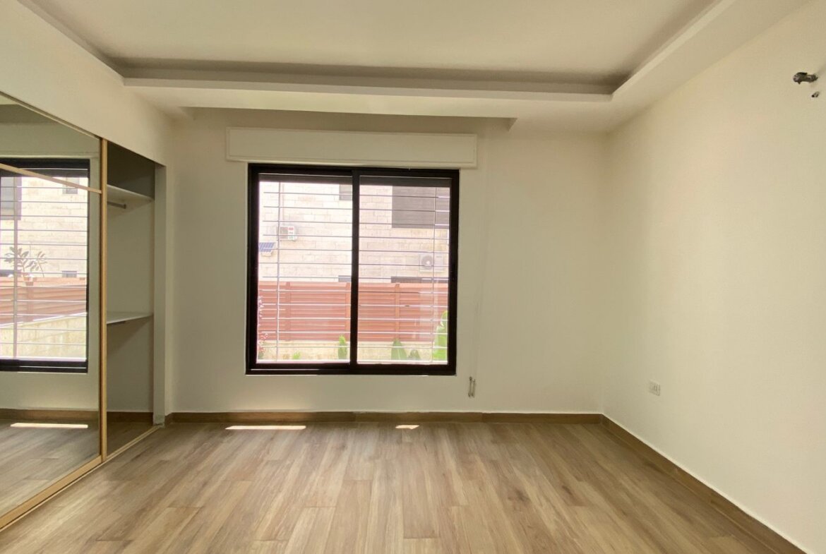 Empty modern room with light wood flooring, large window with blinds on the far wall, and a mirrored sliding-door wardrobe along the left wall; white walls and ceiling with a recessed beam detail.