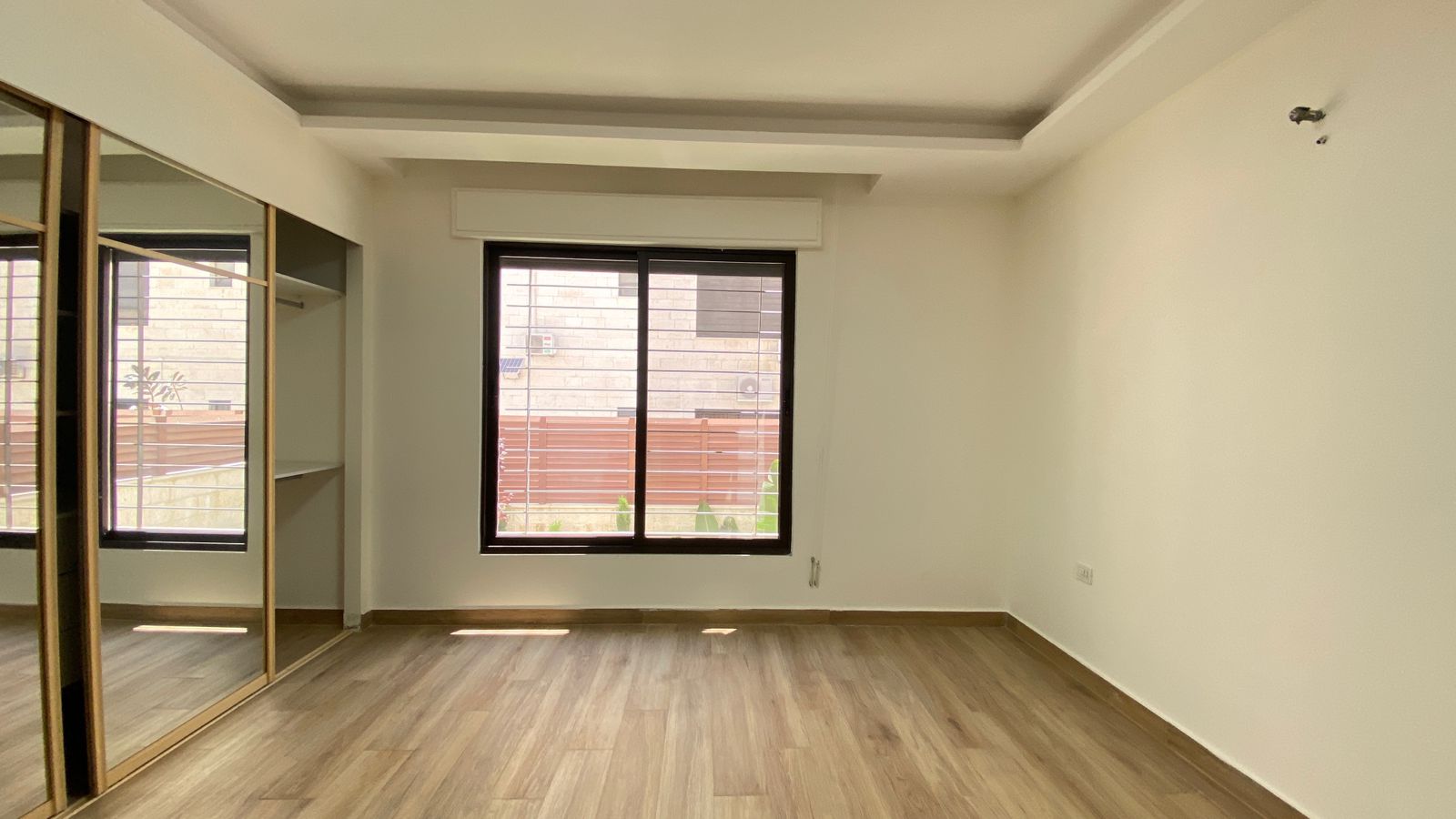 971ebf75-cb6c-4096-bbe1-78a2ad773a21 Empty modern room with light wood flooring, large window with blinds on the far wall, and a mirrored sliding-door wardrobe along the left wall; white walls and ceiling with a recessed beam detail.