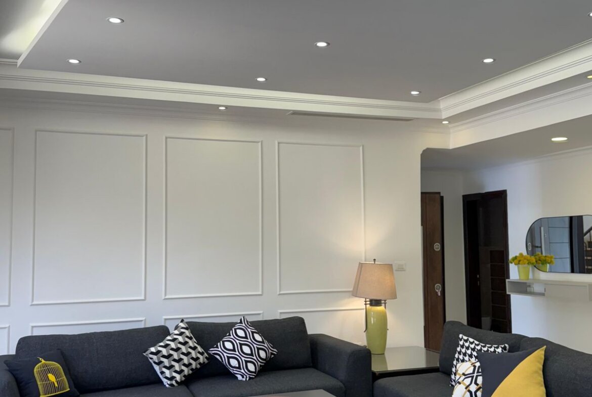Modern living room with dark gray sectional sofa, patterned cushions, and matching ottomans; white paneled walls and recessed ceiling lights.