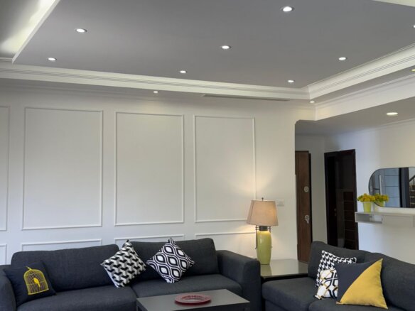 Modern living room with dark gray sectional sofa, patterned cushions, and matching ottomans; white paneled walls and recessed ceiling lights.