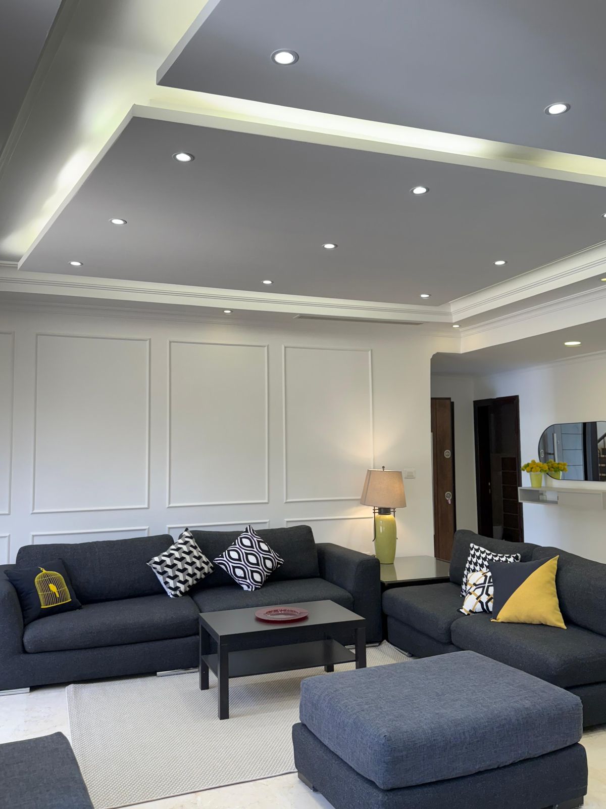 Modern living room with dark gray sectional sofa, patterned cushions, and matching ottomans; white paneled walls and recessed ceiling lights.