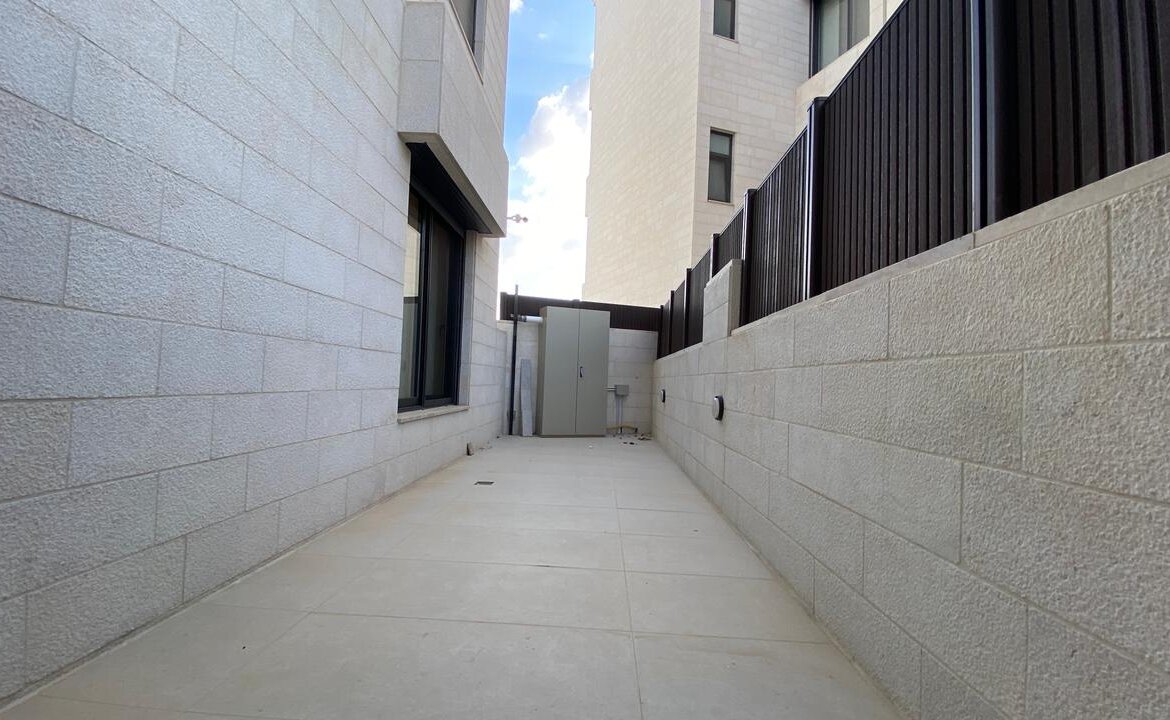 Narrow outdoor concrete corridor between light-gray block buildings, with a tall utility cabinet at the far end and a black metal fence along the right wall; blue sky visible above.