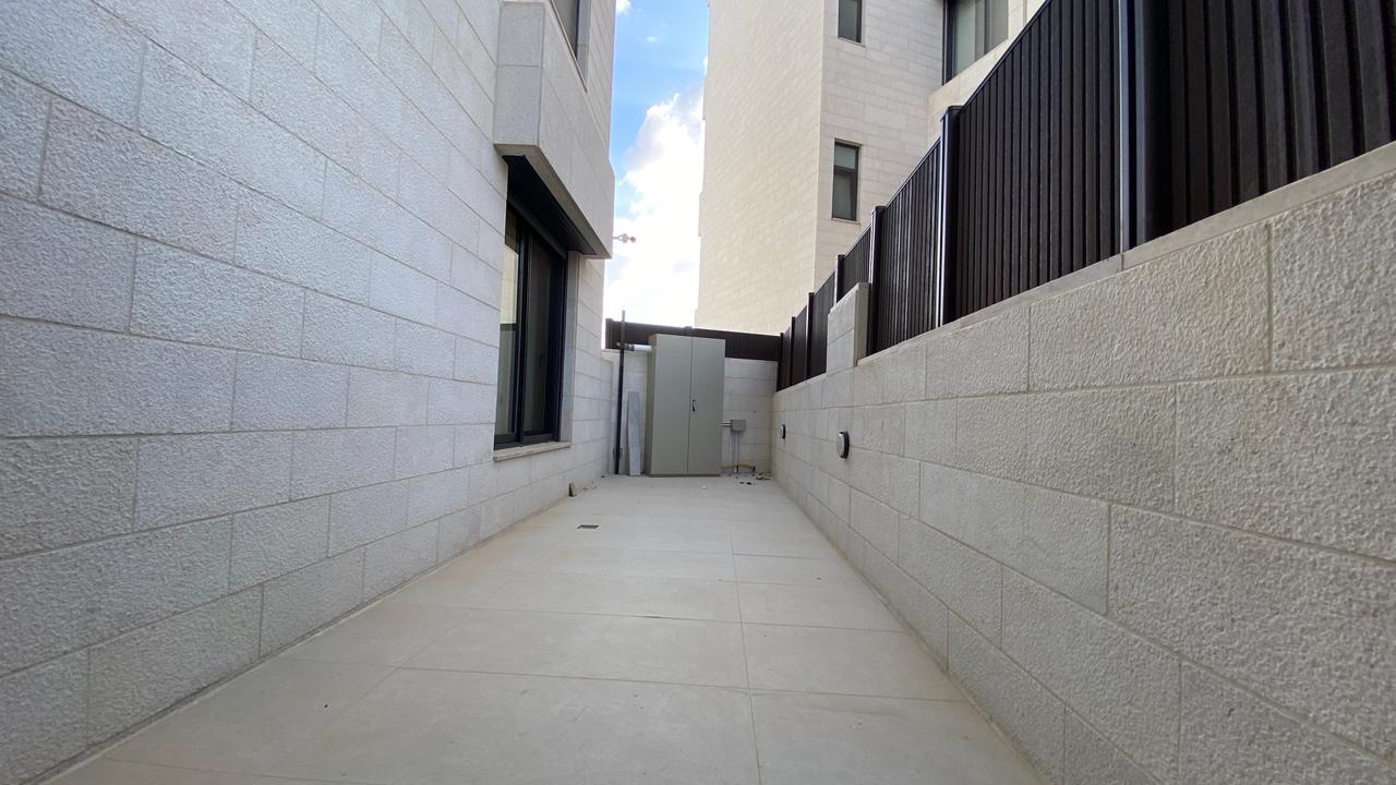 Narrow outdoor concrete corridor between light-gray block buildings, with a tall utility cabinet at the far end and a black metal fence along the right wall; blue sky visible above.