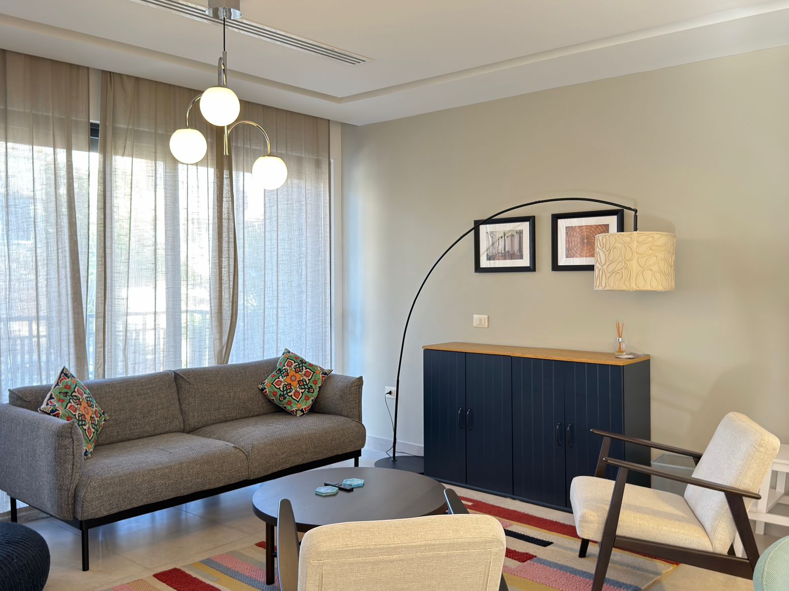 988e4c89-f710-42b5-b41b-35197eacbc99 Cozy living room with a gray sofa, colorful patterned cushions, a round coffee table, and an arc floor lamp over a navy cabinet.