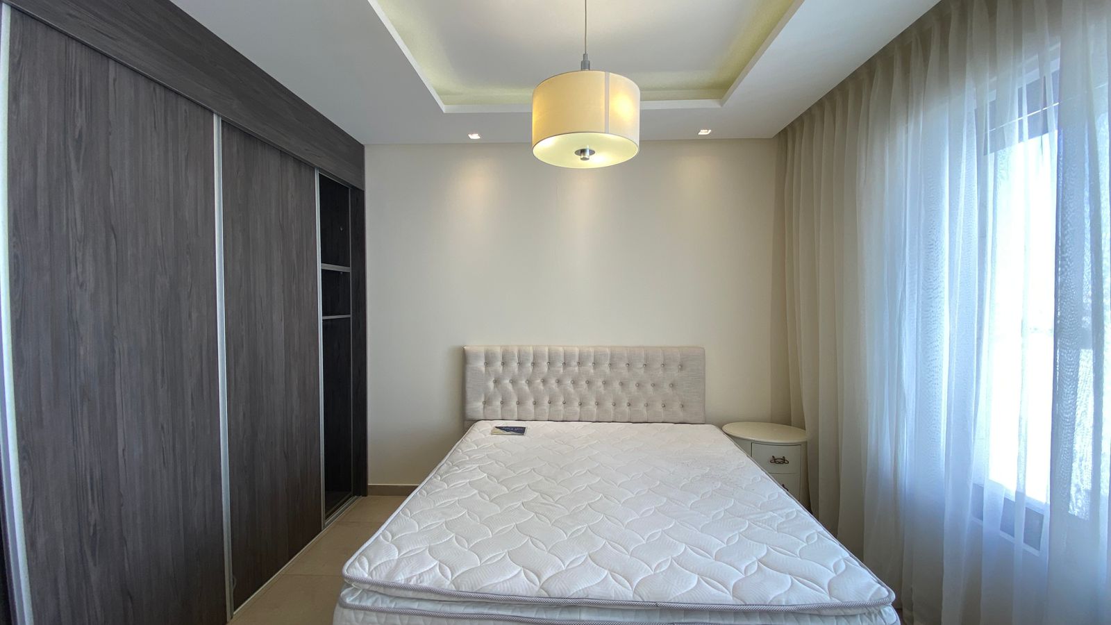 99157677-dd47-45fd-ba07-9d20ad262f73 Modern bedroom with dark wood wardrobes along the left wall, a tufted beige headboard bed in the center, and sheer curtains by a large window on the right.