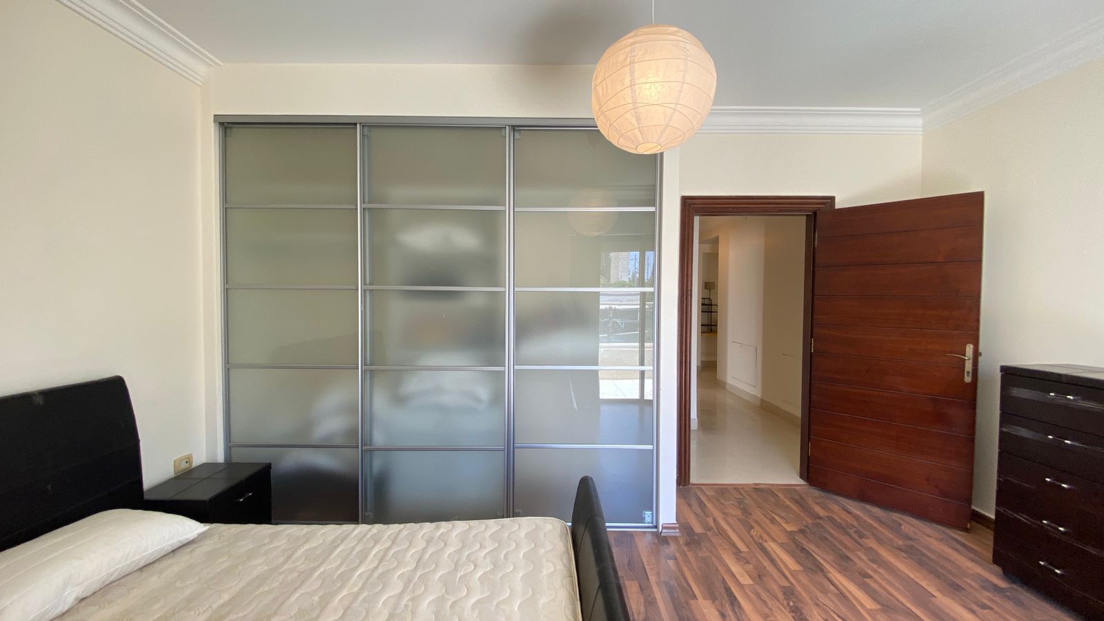 9983d309-1ef2-425b-bafa-db1761ab054c Modern bedroom featuring a frosted-glass sliding wardrobe, a bed with a white quilt, a dark wood nightstand, and a dark wood dresser; an open wooden door leads to a bright hallway, with a round paper lantern hanging from the ceiling and wood laminate flooring.