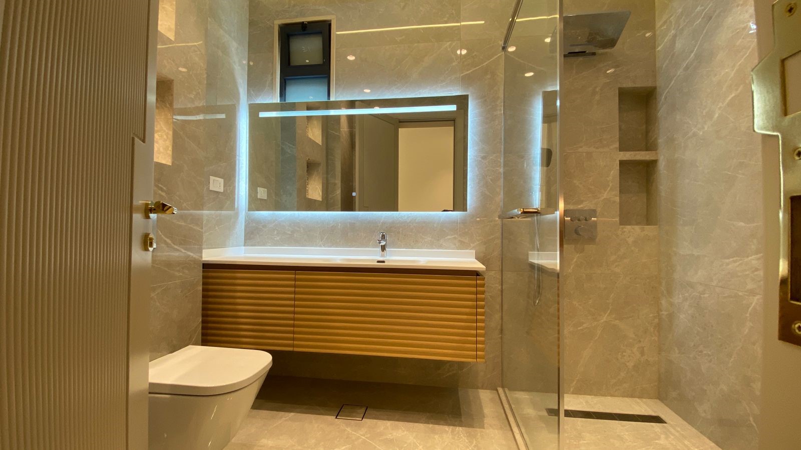 9a0ceb6c-711e-4f70-812c-65d42221abf0 Modern marble bathroom with a wall-mounted wooden vanity, backlit mirror, and a glass shower enclosure beside a white toilet.