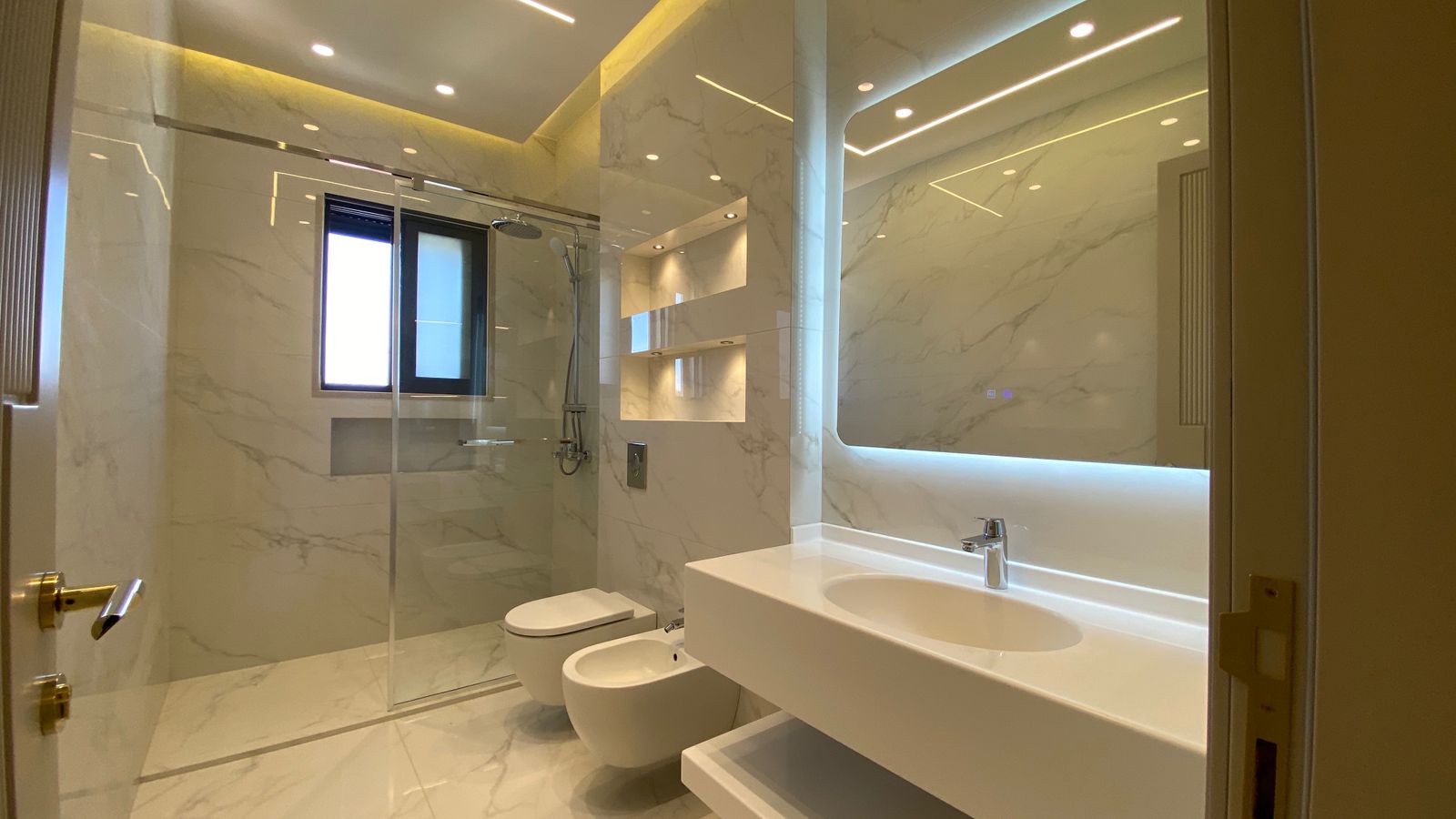 9af52867-50e8-4fa7-911b-3680daa0002c Modern marble bathroom with glass-enclosed shower, wall-mounted sink, bidet, and backlit mirror.