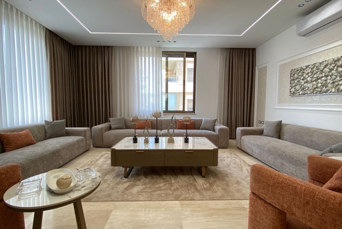 Luxurious living room with three gray sectionals, a marble coffee table, and a crystal chandelier above a neutral rug.